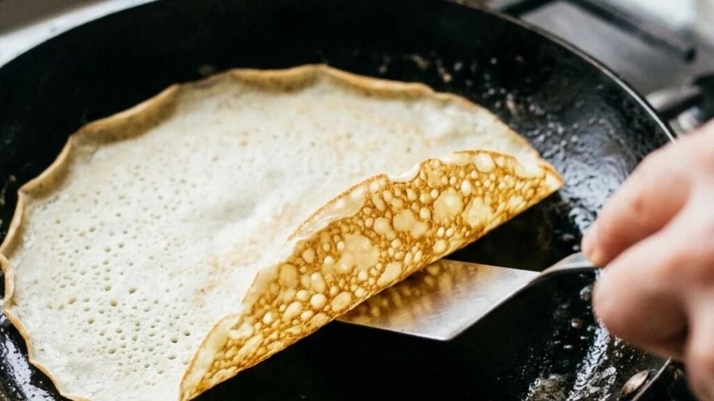 A spatula lifting the lightly golden edge of a thin pancake in a frying pan.